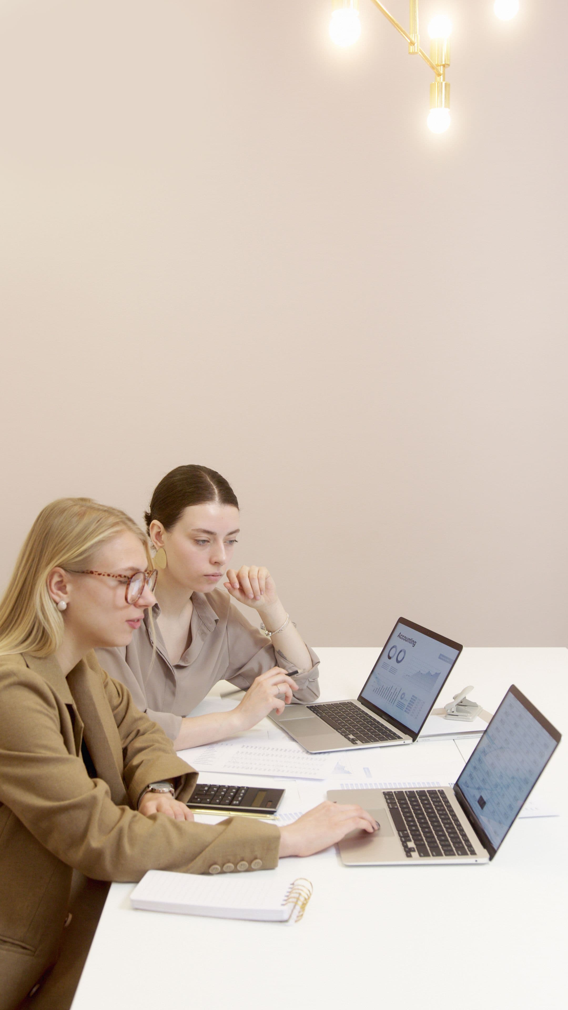 Two professional women analyze financial data on laptops at a white office desk.