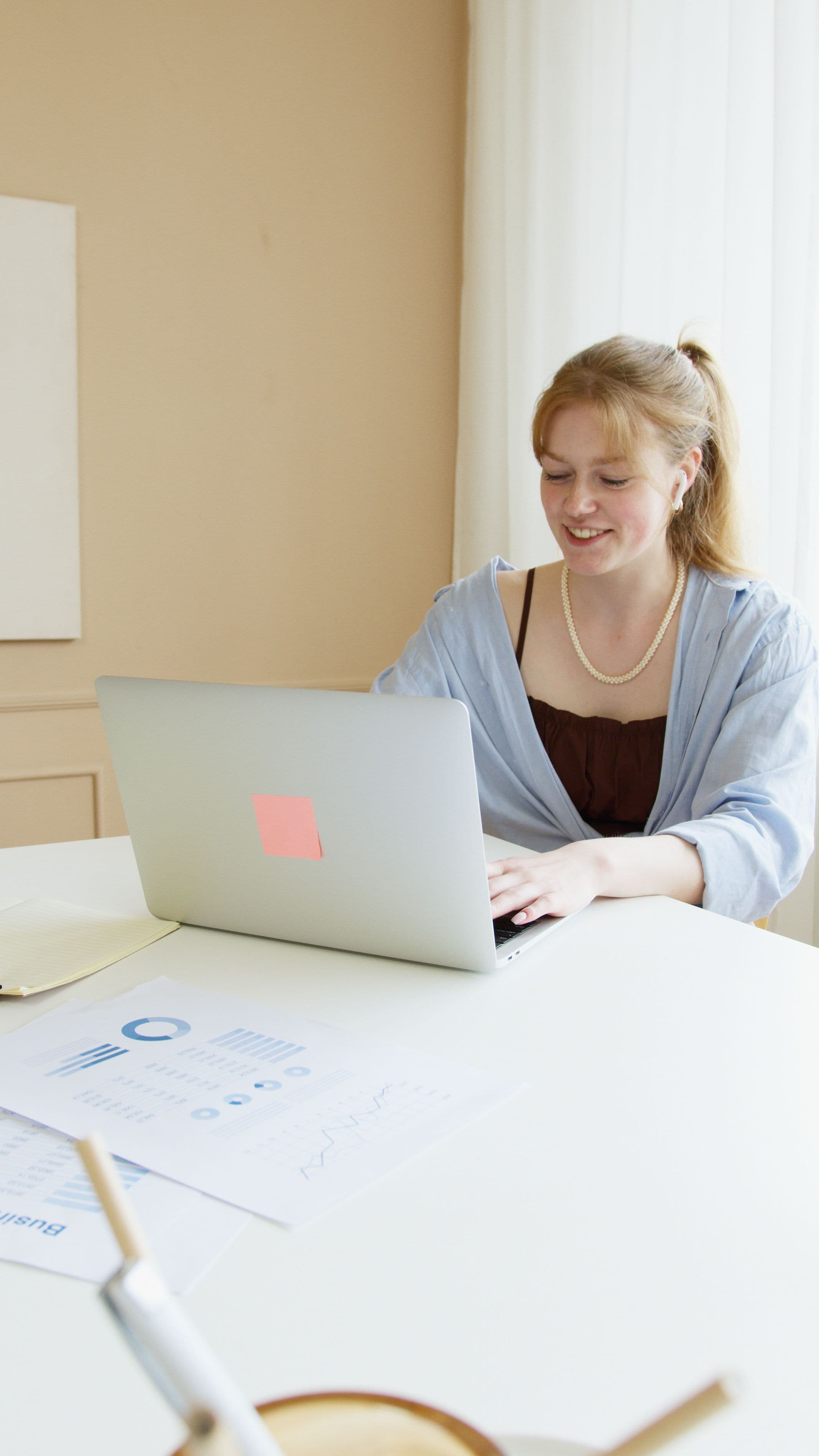 Smiling woman with red hair using a laptop at a white desk with data charts.
