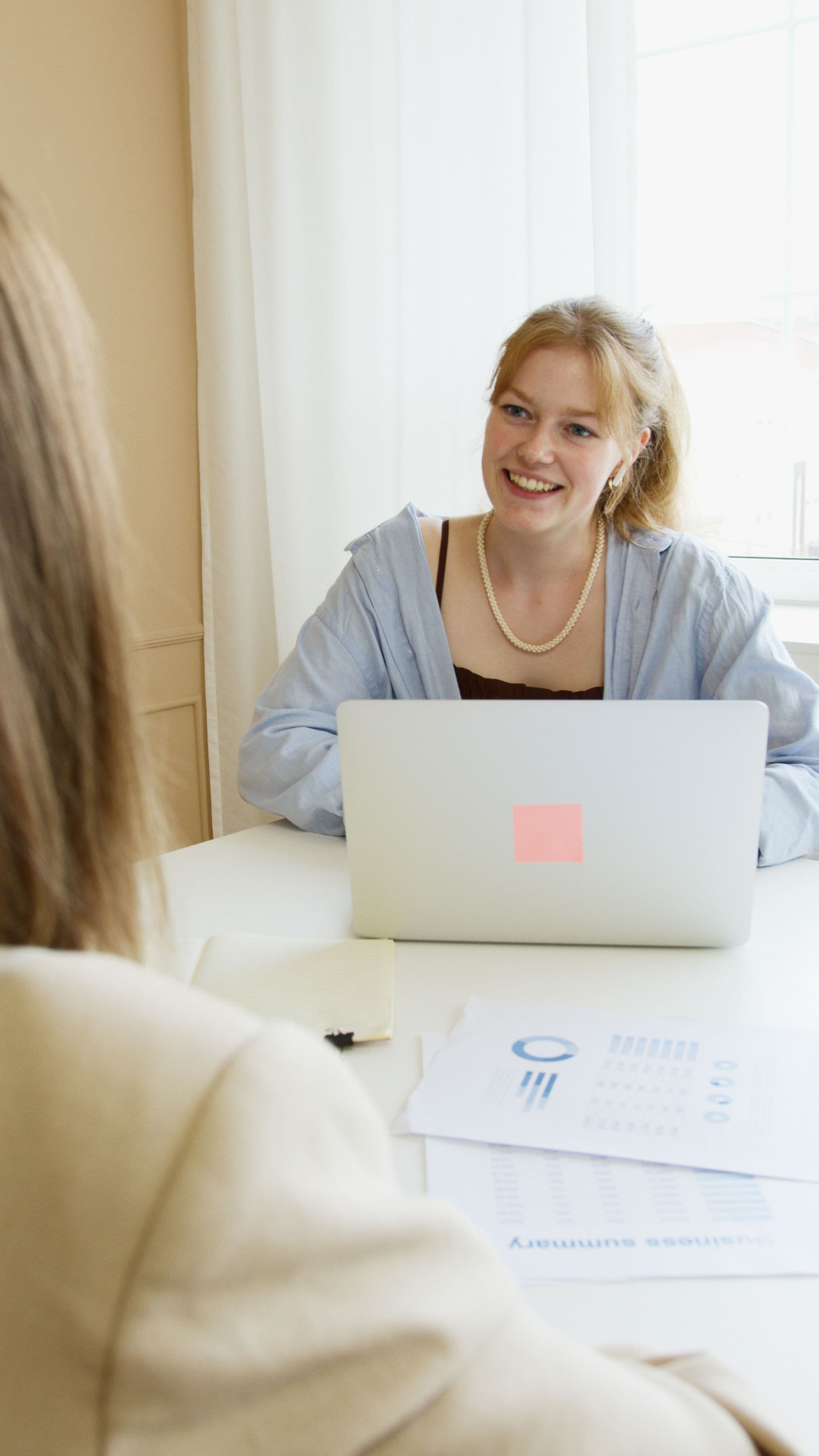 Smiling woman sits at a white desk with a laptop and business documents.
