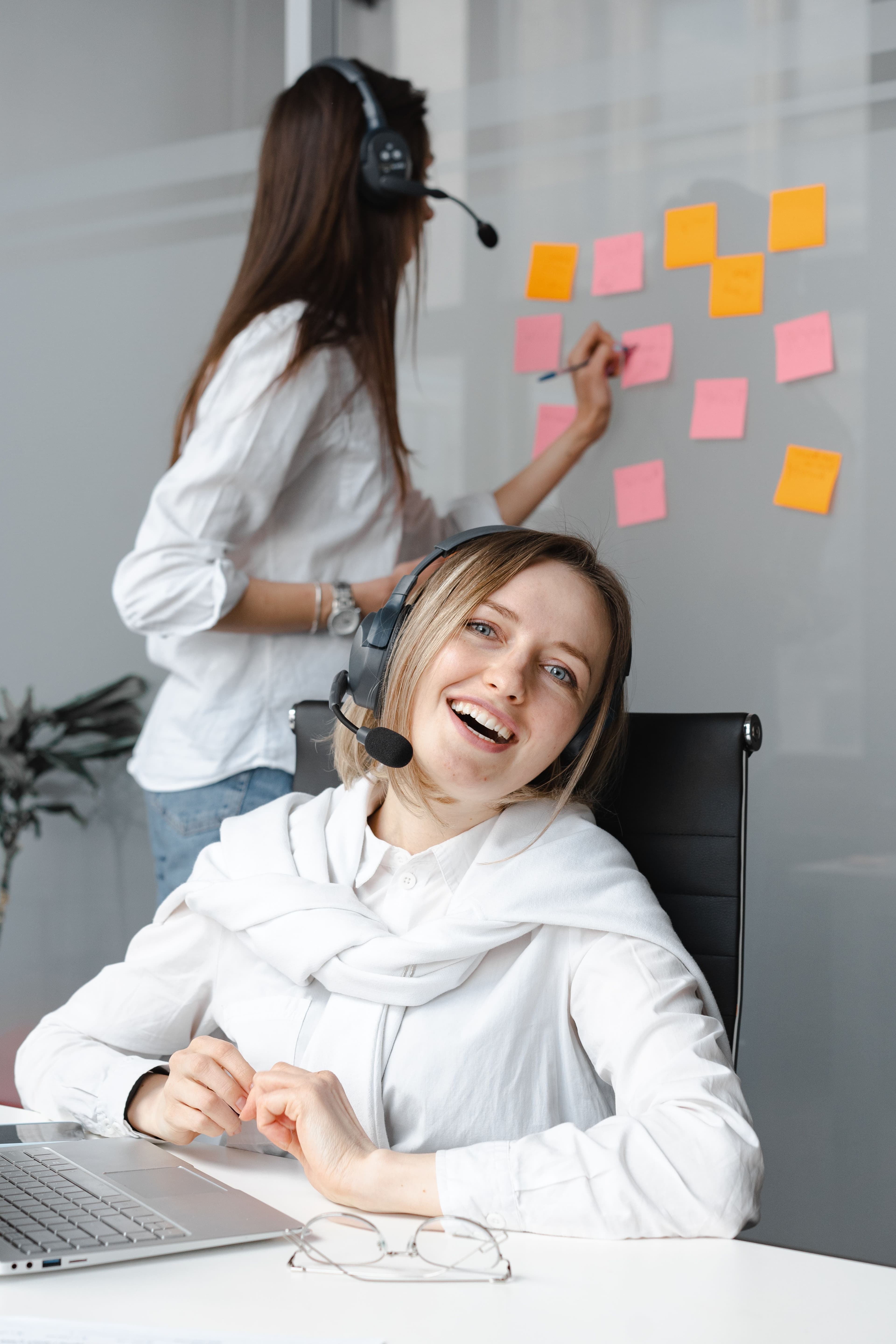 Smiling woman wearing a headset at a desk while a colleague writes on colorful sticky notes.