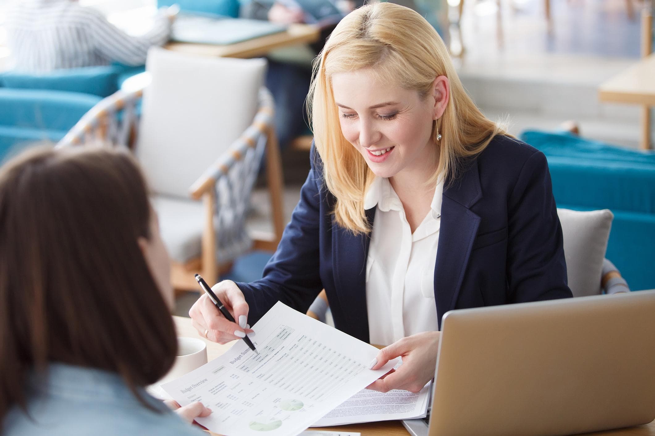 Smiling blonde woman in a navy blazer reviews a budget document with a colleague.